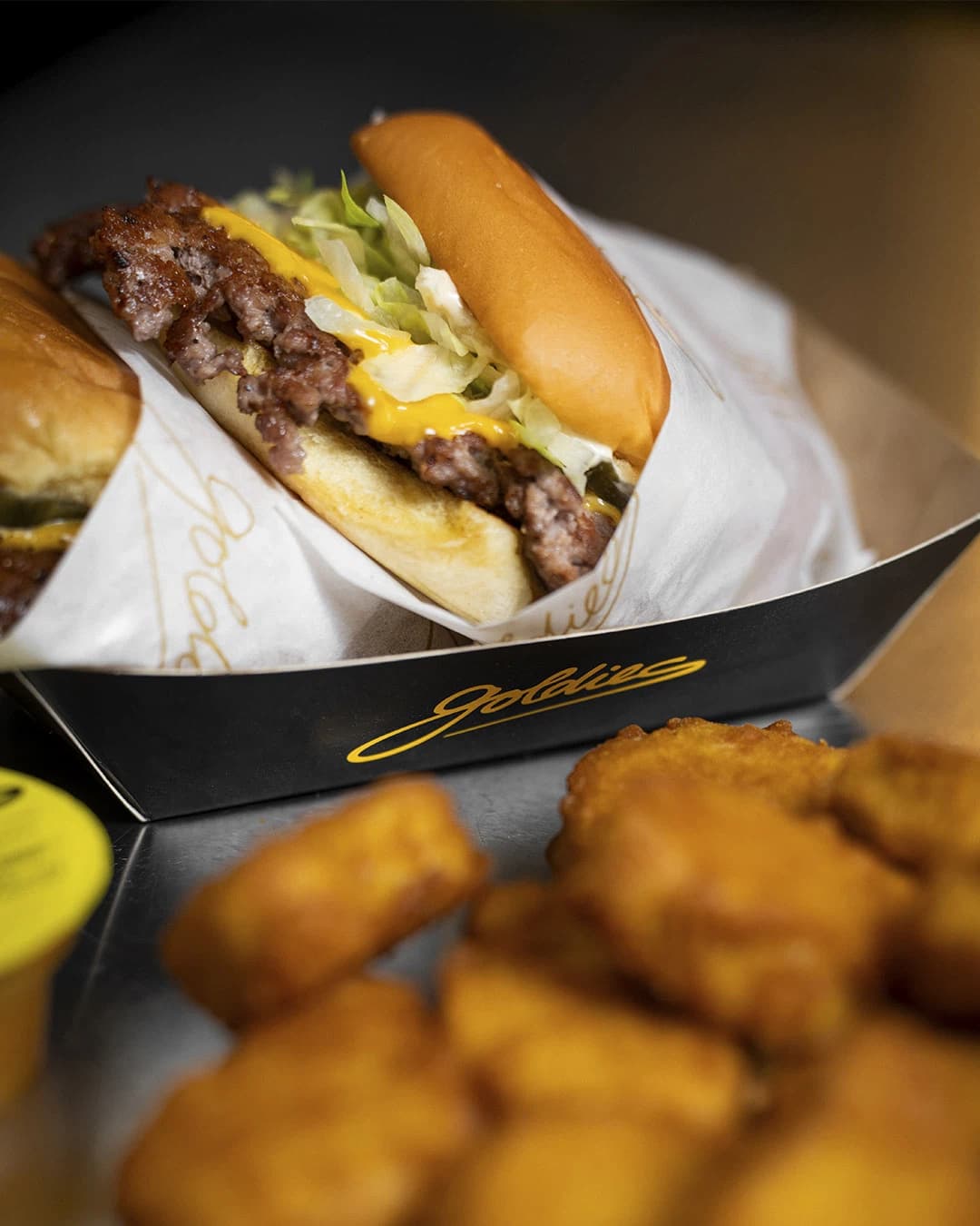 Goldies Smashburger mit Käse in der Papierschale, daneben Chicken Nuggets